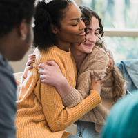 photo of white female high school student and black female high school student sitting in a circle of chairs hugging while a black male high school student looks on